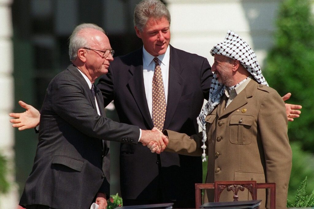 As President Bill Clinton smiles, Israeli Prime Minister Yitzhak Rabin and PLO Chairman Yasser Arafat shake hands on the White House lawn, on Sept. 13, 1993. After months of secret negotiations in Norway, Israeli and Palestinian negotiators produced the Oslo accords, a land-for-peace deal that established the Palestinian Authority in the West Bank and Gaza, began an Israeli withdrawal from occupied territories, and called for talks on the conflict's core issues—including Palestinian refugees, Israeli settlements, and Jerusalem—after a five-year interim period.