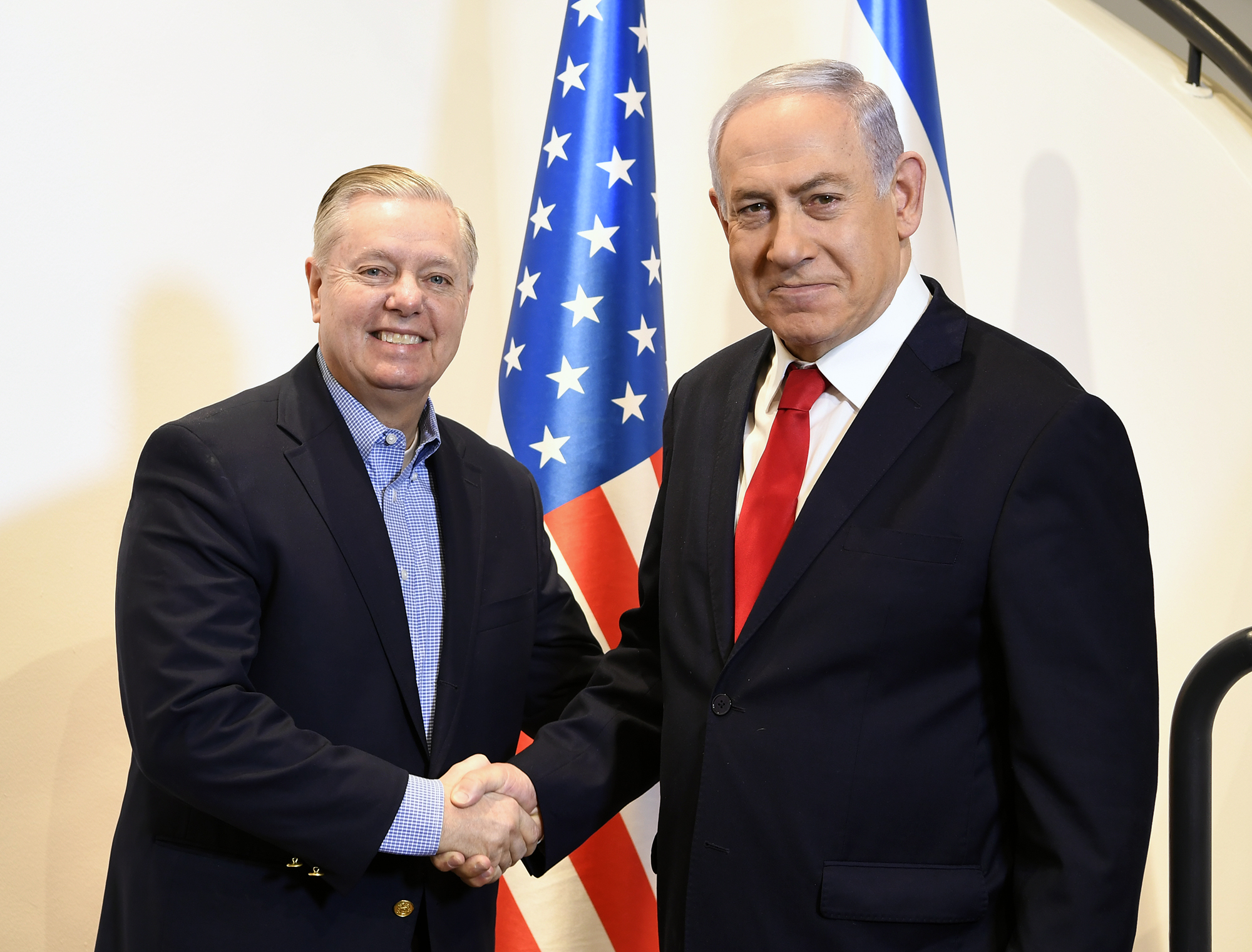 Two men in dark suits shake hands in front of a U.S. flag, posing for a formal photo op.